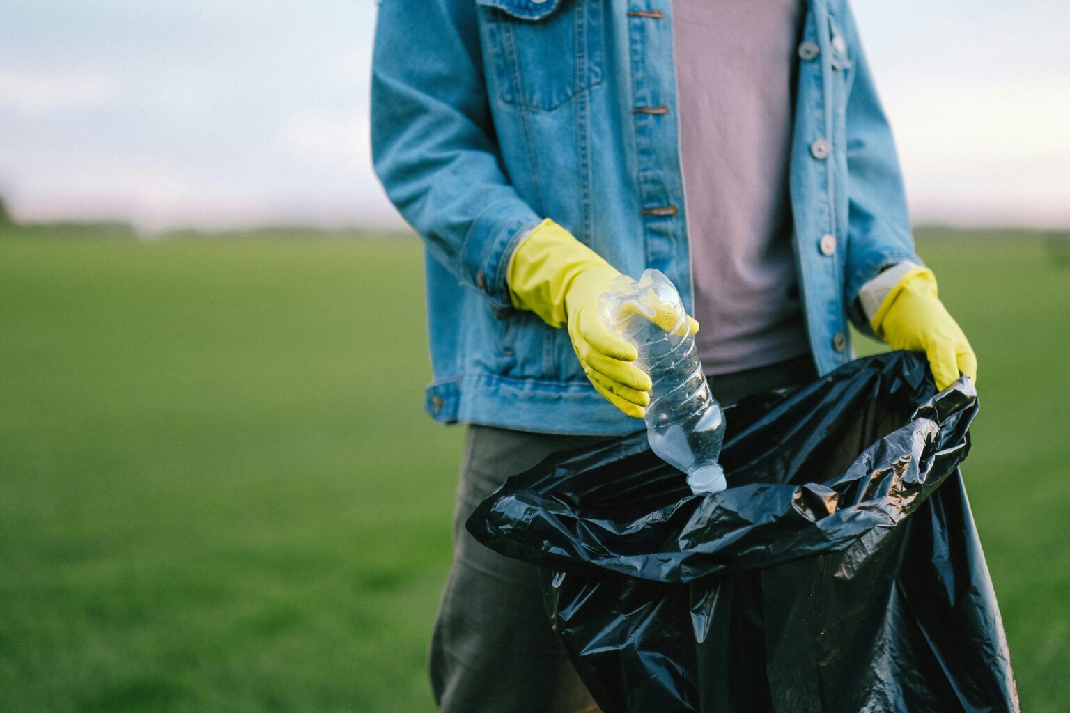 Eine Person, die gelbe Handschuhe und eine Jeansjacke trägt, hebt eine Plastikflasche auf und steckt sie in einen schwarzen Müllsack auf einer Wiese im Freien.