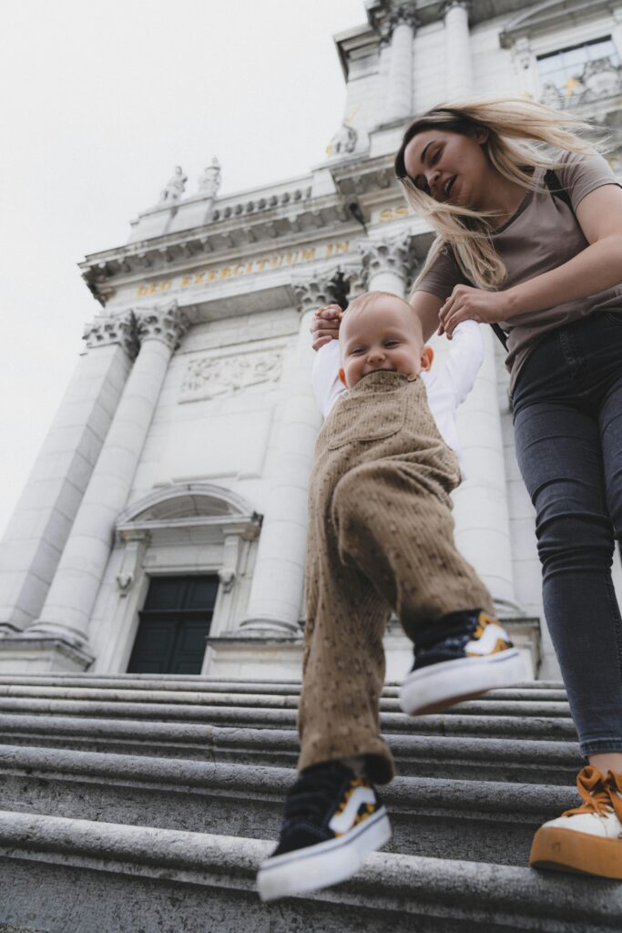 Eine Frau hilft einem lächelnden Kleinkind in einem braunen Overall die Steinstufen vor einem großen, weißen Gebäude mit Säulen hinunter.