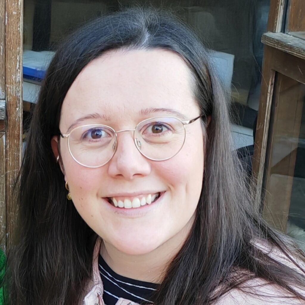 Eine Person mit langen braunen Haaren, einer runden Brille und einer hellrosa Jacke lächelt in die Kamera, die draußen an einem Holzfenster steht.