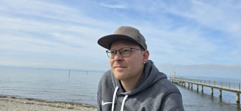 Ein Mann mit Brille und Mütze und grauem Kapuzenpulli steht in der Nähe eines ruhigen Strandes mit einem ins Wasser ragenden Steg unter einem blauen Wolkenhimmel.