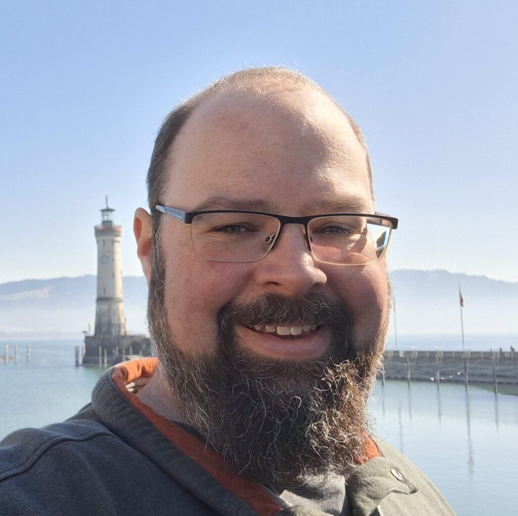 Ein bärtiger Mann mit Brille lächelt an einem klaren, sonnigen Tag vor einem Leuchtturm am Wasser. Im Hintergrund sind Berge und eine Seebrücke zu sehen.