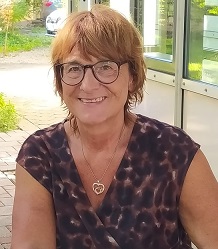 Eine lächelnde Frau mit kurzen braunen Haaren und Brille sitzt im Freien, trägt ein gemustertes Kleid und eine herzförmige Halskette.