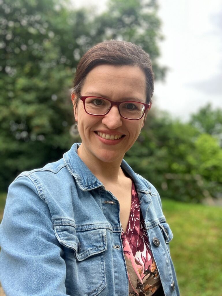Eine Frau mit Brille und Jeansjacke lächelt im Freien, mit grünen Bäumen und Gras im unscharfen Hintergrund.