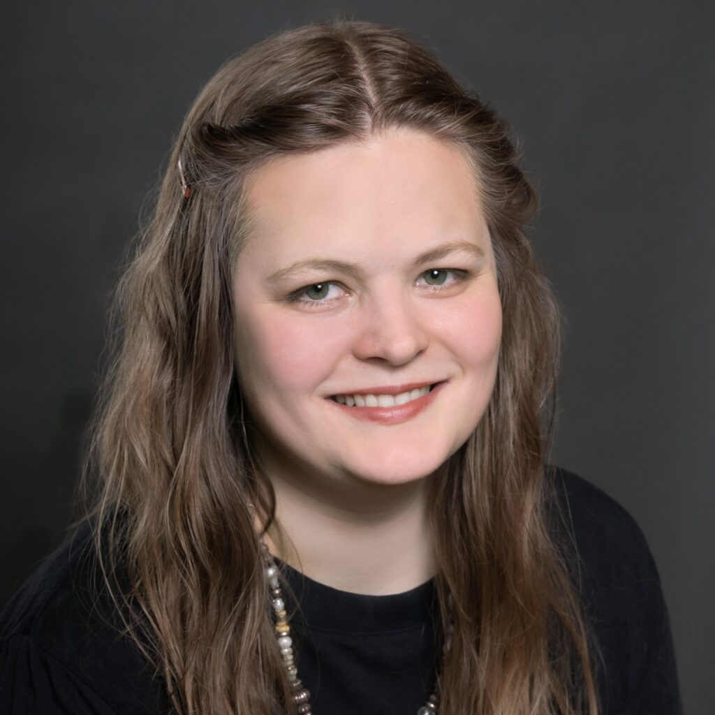 Eine Frau mit langen, gewellten braunen Haaren lächelt in die Kamera. Sie trägt ein schwarzes Oberteil und eine Perlenkette, dahinter ist ein dunkelgrauer Hintergrund zu sehen.