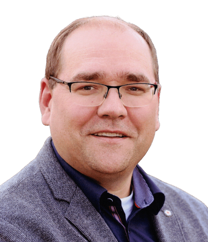 Ein lächelnder Mann mit kurzem Haar und Brille, der einen grauen Blazer über einem dunklen Hemd trägt, posiert vor einem schlichten weißen Hintergrund.