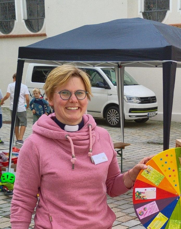 Eine lächelnde Person in einem rosa Kapuzenpulli und mit klerikalem Kragen hält ein buntes Spinnrad bei einer Veranstaltung im Freien, mit einem weißen Lieferwagen und Kindern im Hintergrund.