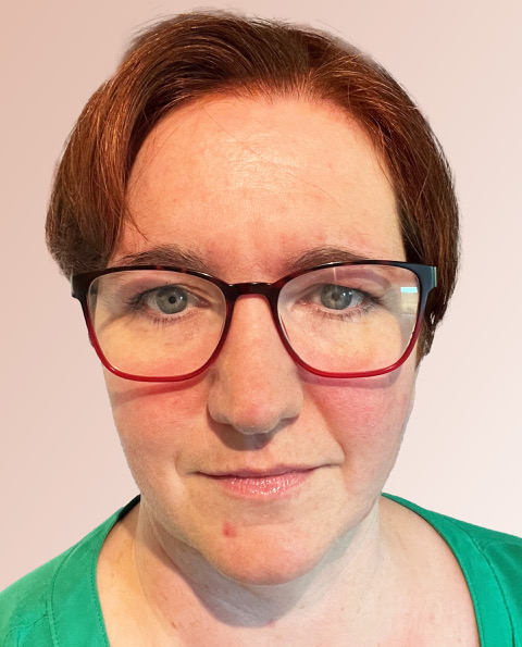 Eine Person mit kurzen braunen Haaren, einer rotgeränderten Brille und einem grünen Oberteil blickt direkt in die Kamera vor einem hellrosa Hintergrund.