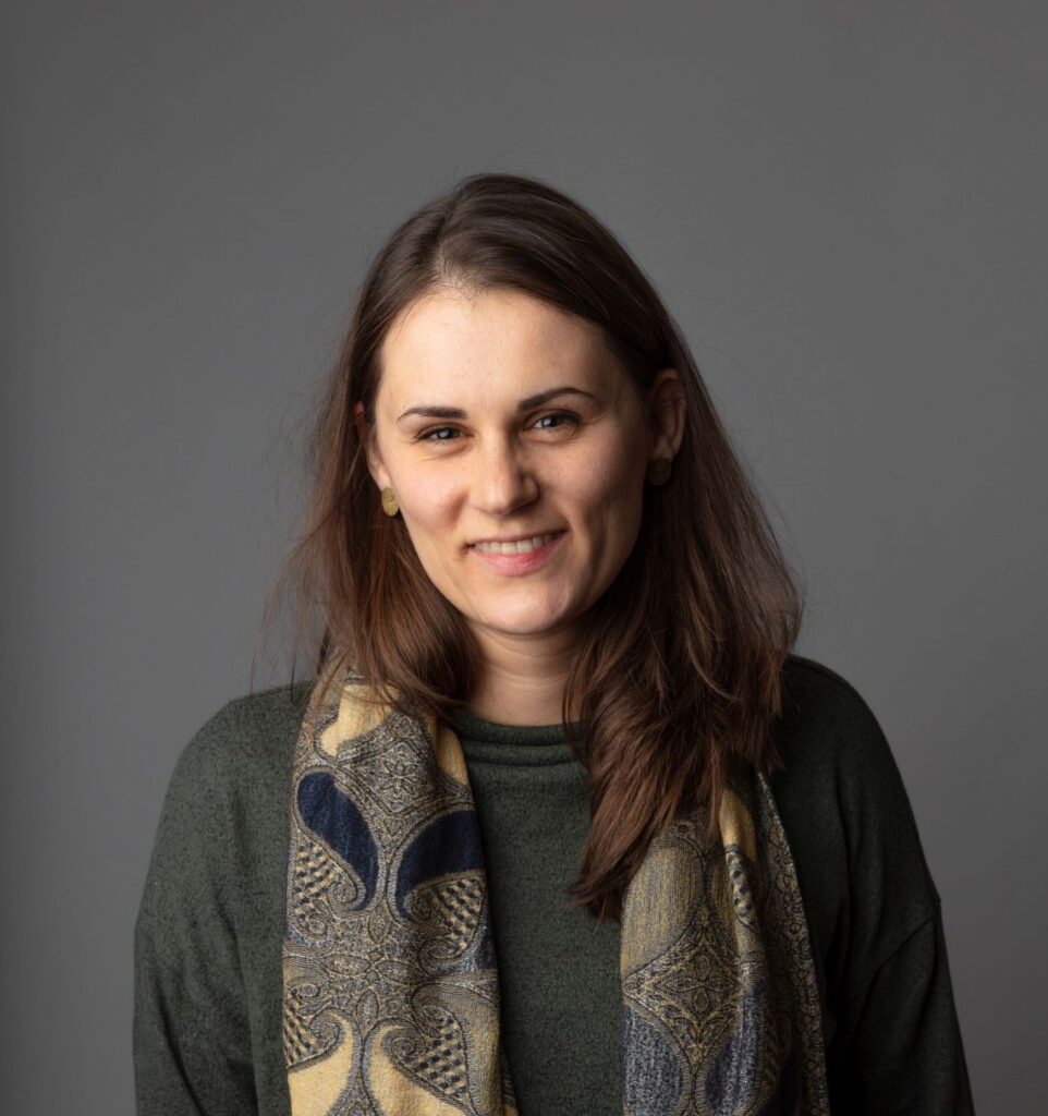 Eine Frau mit langen braunen Haaren, die einen grünen Pullover und einen gemusterten Schal trägt, lächelt vor einem schlichten grauen Hintergrund.