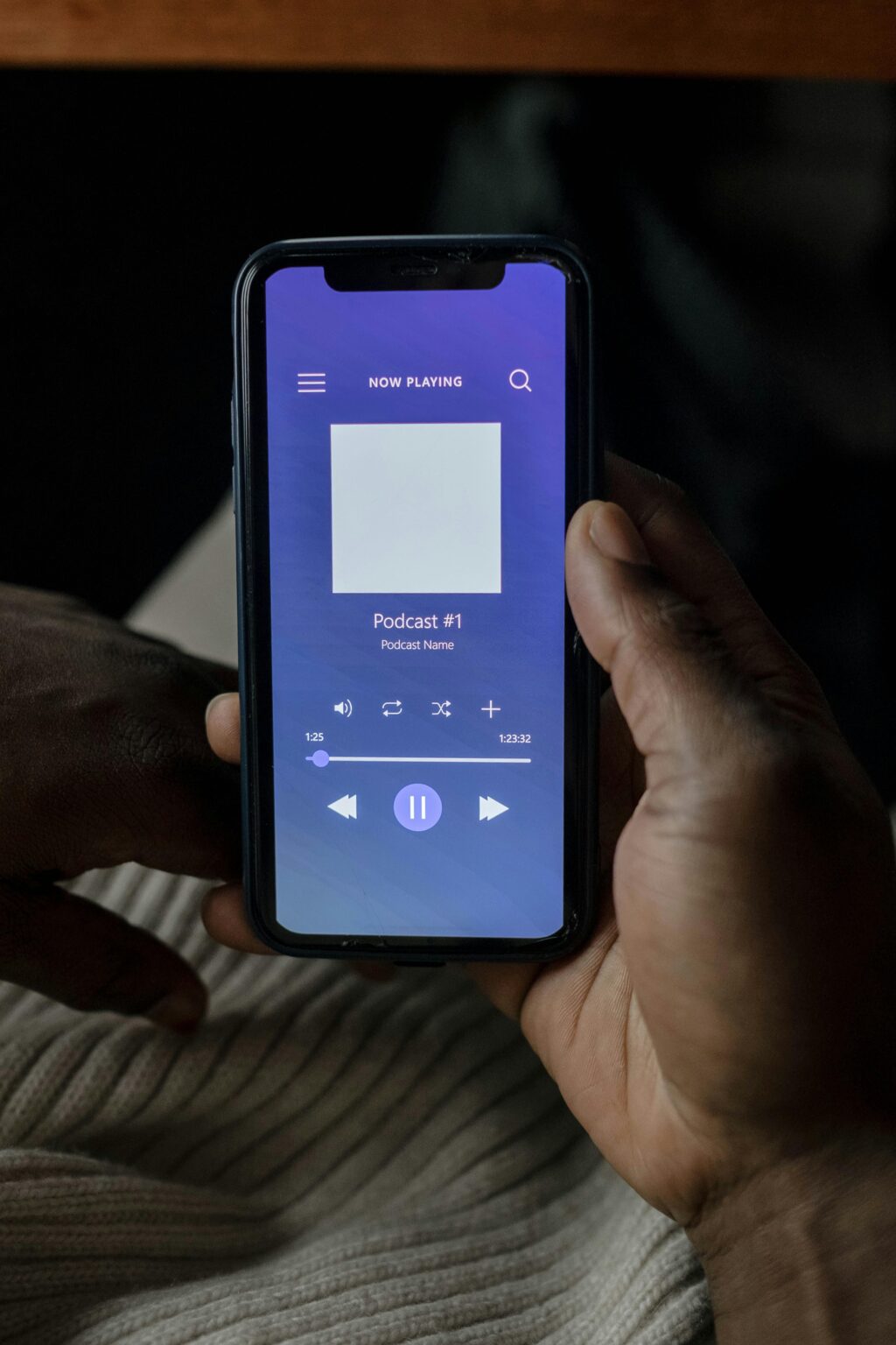 Eine Person hält ein Smartphone in der Hand, auf dem der Bildschirm eines Podcast-Players mit Wiedergabesteuerungen und "Podcast Nr. 1" als aktuelle Wiedergabe angezeigt wird.
