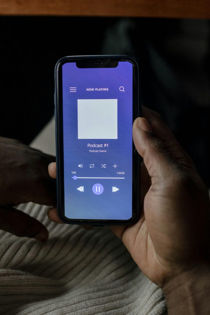 Eine Person hält ein Smartphone in der Hand, auf dem der Bildschirm eines Podcast-Players mit Wiedergabesteuerungen und "Podcast Nr. 1" als aktuelle Wiedergabe angezeigt wird.