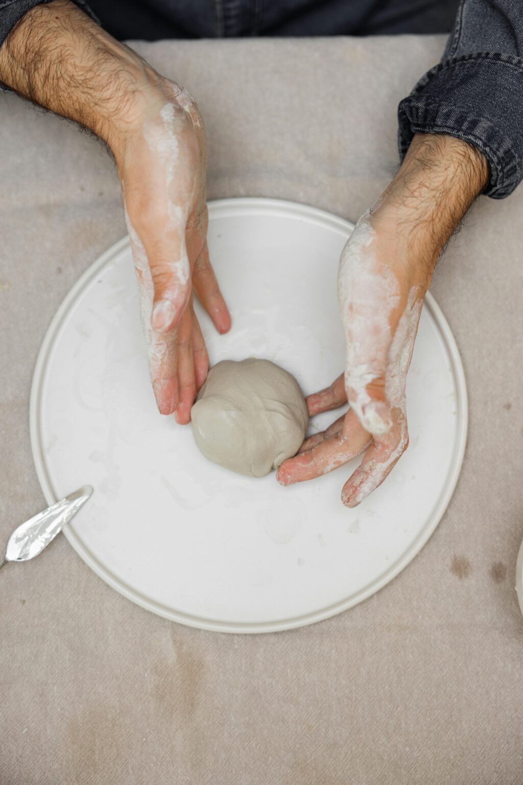 Eine Person mit tonbedeckten Händen formt eine Tonkugel auf einer weißen Platte und bereitet sich so auf die Töpfer- oder Bildhauerarbeit vor.
