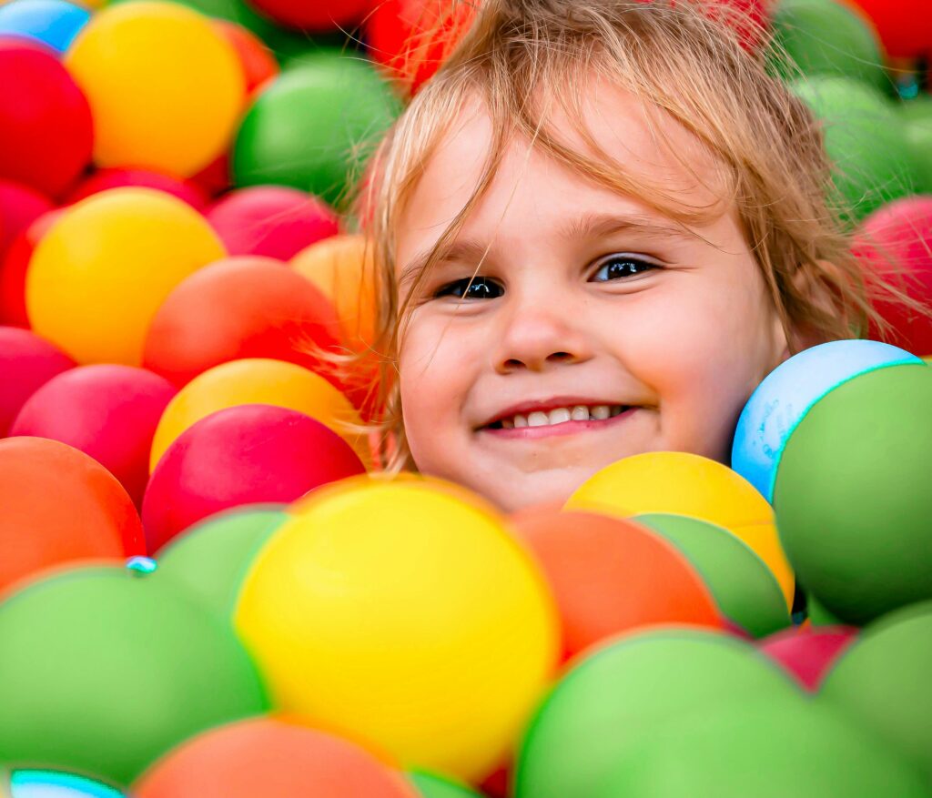 Lächelndes Kleinkind, umgeben von bunten Plastikbällen in einer Ballgrube, dessen Gesicht zwischen den roten, gelben, grünen und orangefarbenen Bällen teilweise sichtbar ist.