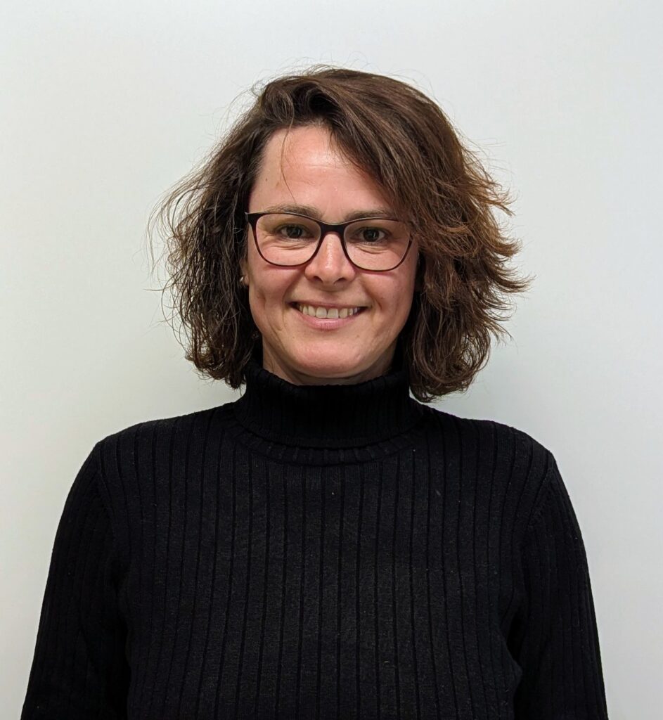 Eine Frau mit gewelltem braunem Haar und Brille lächelt in die Kamera. Sie trägt einen schwarzen gerippten Rollkragenpullover vor einem schlichten weißen Hintergrund.
