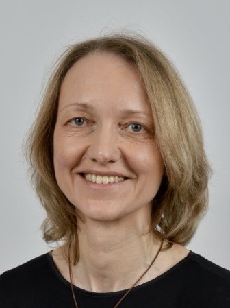 Eine Frau mit schulterlangem blondem Haar, die ein schwarzes Oberteil trägt und lächelt, posiert vor einem schlichten hellgrauen Hintergrund.