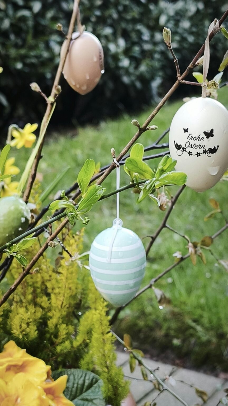Dekorative Ostereier hängen an knospenden Zweigen in einem Garten, mit gelben Blumen und üppigem Grün im Hintergrund.