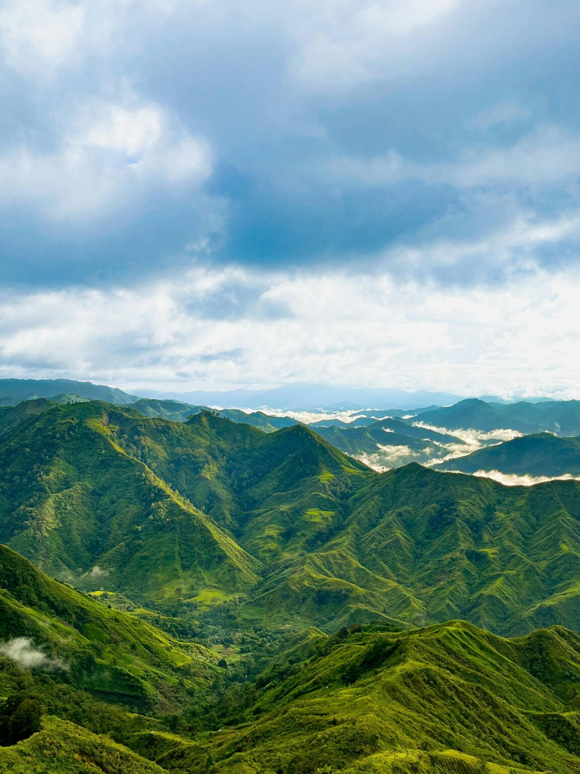 Üppig grüne Berge unter einem teilweise bewölkten Himmel, mit tief liegendem Nebel in den Tälern und Sonnenlicht, das die lebendige Landschaft beleuchtet.
