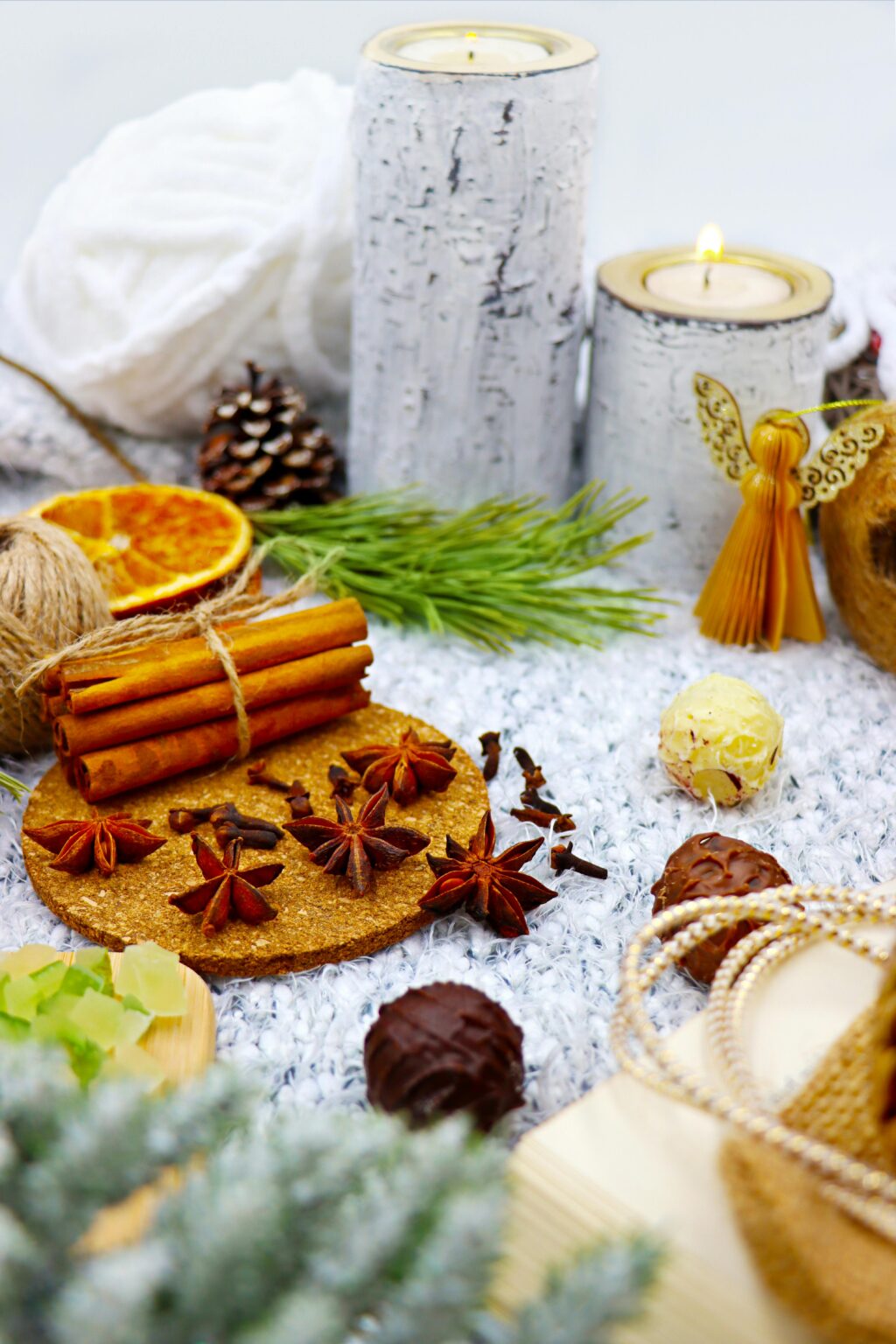 Eine gemütliche Winterszene mit Zimtstangen, Sternanis, getrockneten Orangen, Tannenzapfen, Kerzen, Tannenzweigen, einem Stroh-Engel und festlichen Dekorationen auf einer weichen Oberfläche.