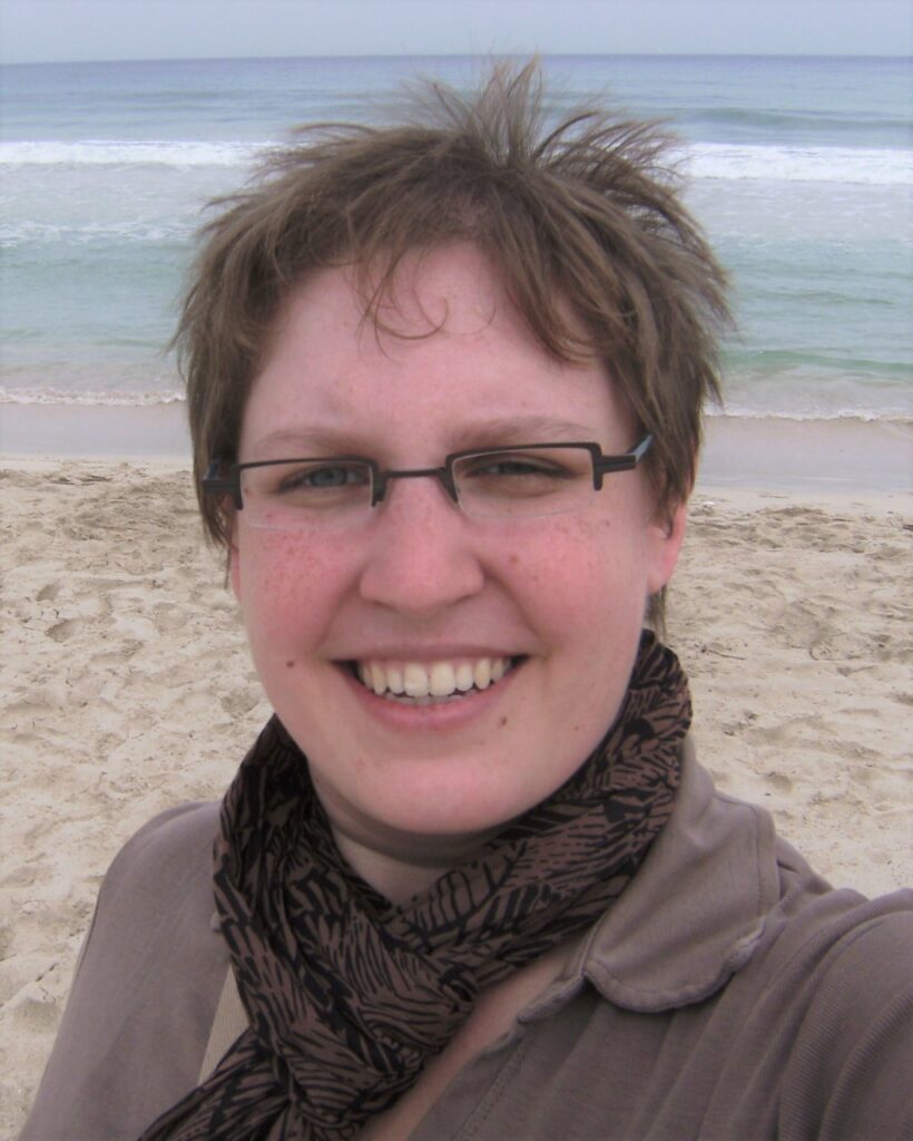 Eine Person, die eine Brille und einen Schal trägt, lächelt an einem Strand mit dem Meer im Hintergrund.