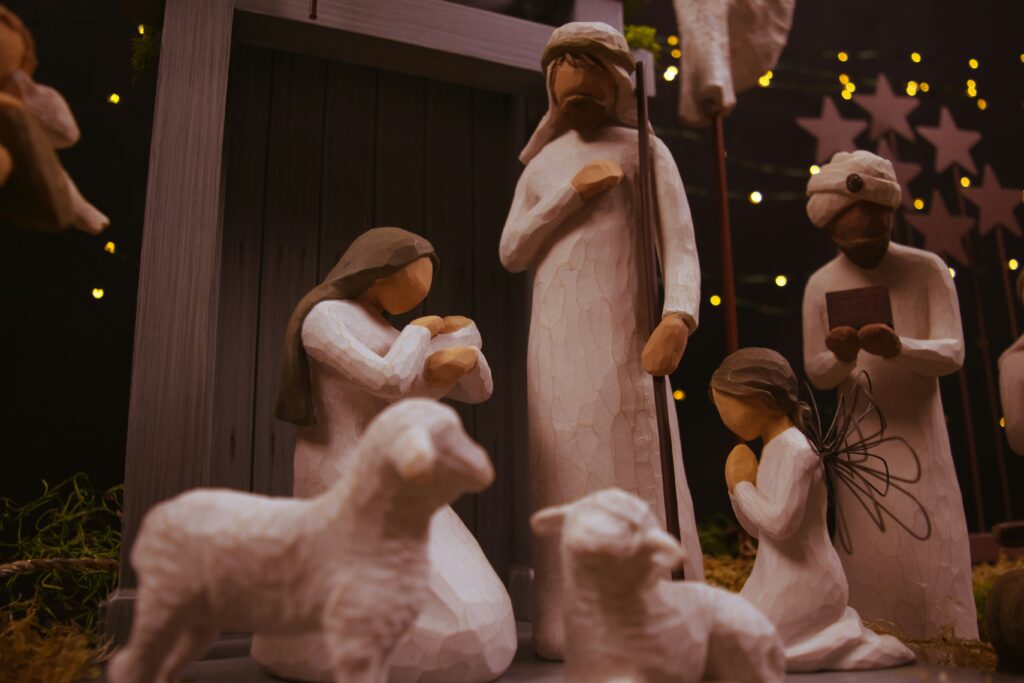 Krippe aus Holz mit Maria, Josef, dem Jesuskind, zwei Schafen, einem Engel und einem weisen Mann vor einer Kulisse aus leuchtenden Lichterketten und Sternen.