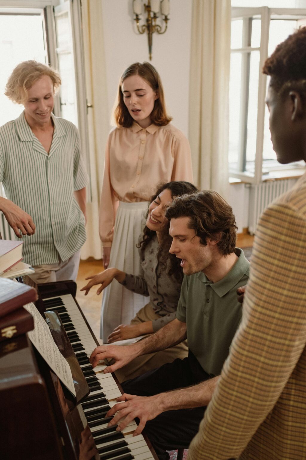 Fünf Personen versammeln sich um ein Klavier, wobei eine Person spielt, während die anderen daneben stehen oder sitzen und in einem hellen, gemütlichen Raum zu singen oder zuzuhören scheinen.