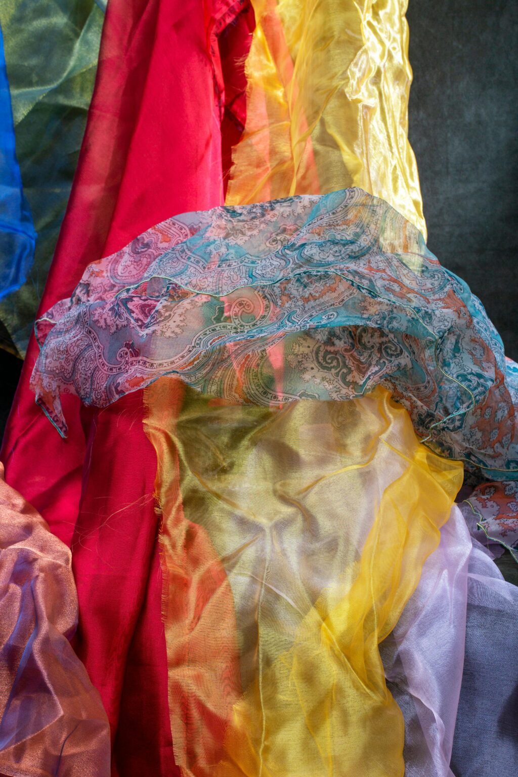 Ein farbenfroher, durchsichtiger Stoff in Rot, Gelb, Violett und Blau, der mit einem blau gemusterten Spitzentextil überlagert ist und so ein lebhaftes, strukturiertes Bild ergibt.