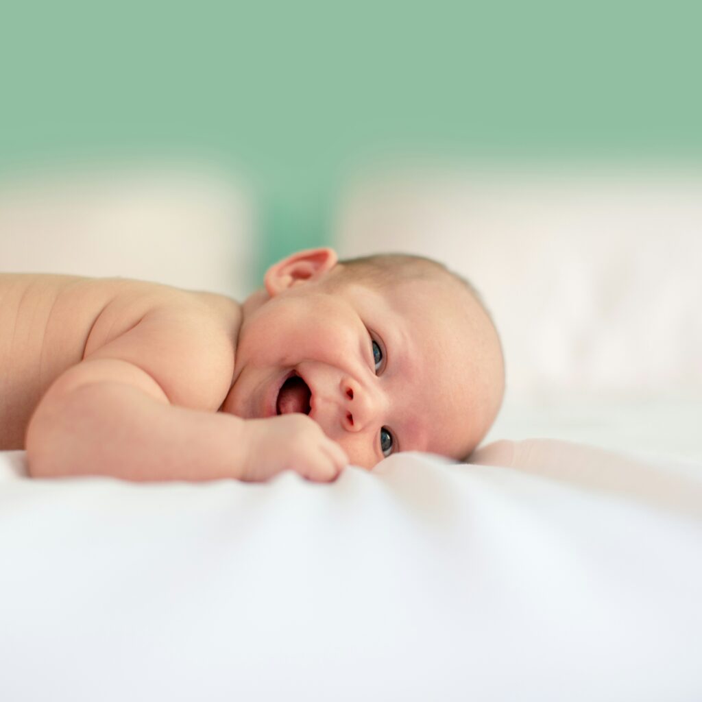 Ein lächelndes Baby liegt auf dem Bauch auf einem weißen Bett und schaut vor einem hellgrünen Hintergrund in die Kamera.